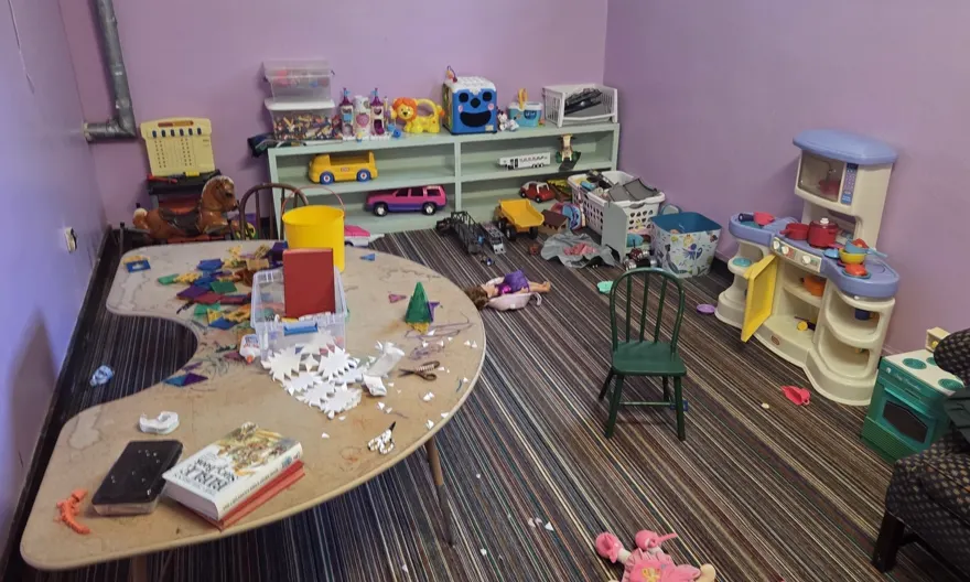 A church nursery before cleaning: toys scattered across the floor and craft debris covering the table.